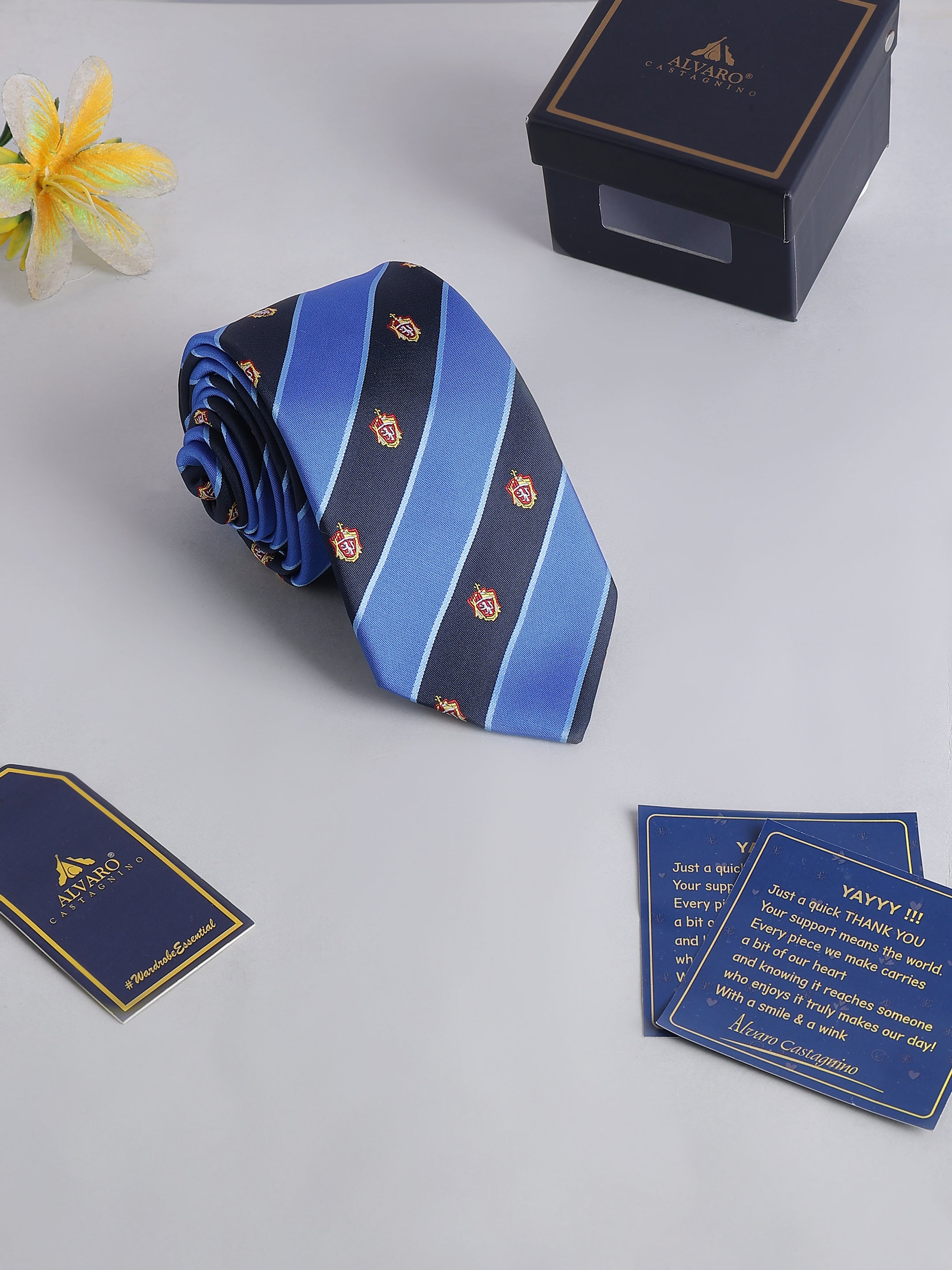 Blue striped tie with decorative elements on a light gray surface, featuring a branded box and card.