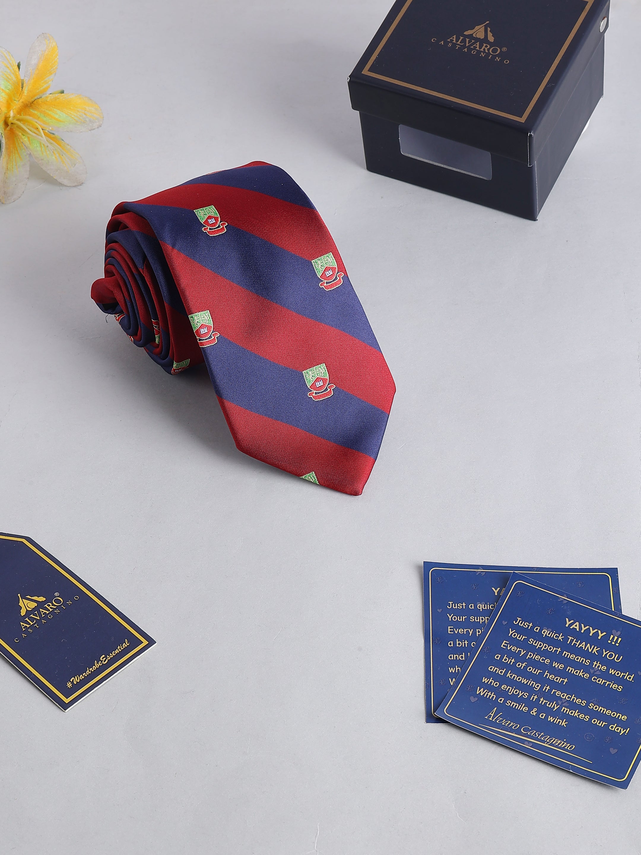 Red and blue striped tie with a brand logo, placed on a light surface with branded cards and box.