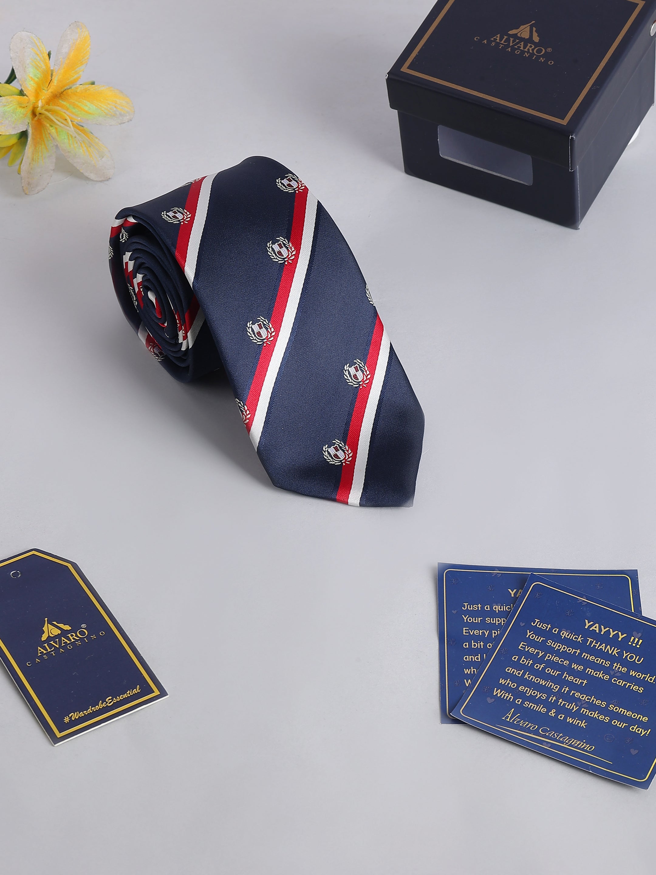 Navy blue tie with red and white stripes and logo, placed on a light gray surface with a box and cards in the background.
