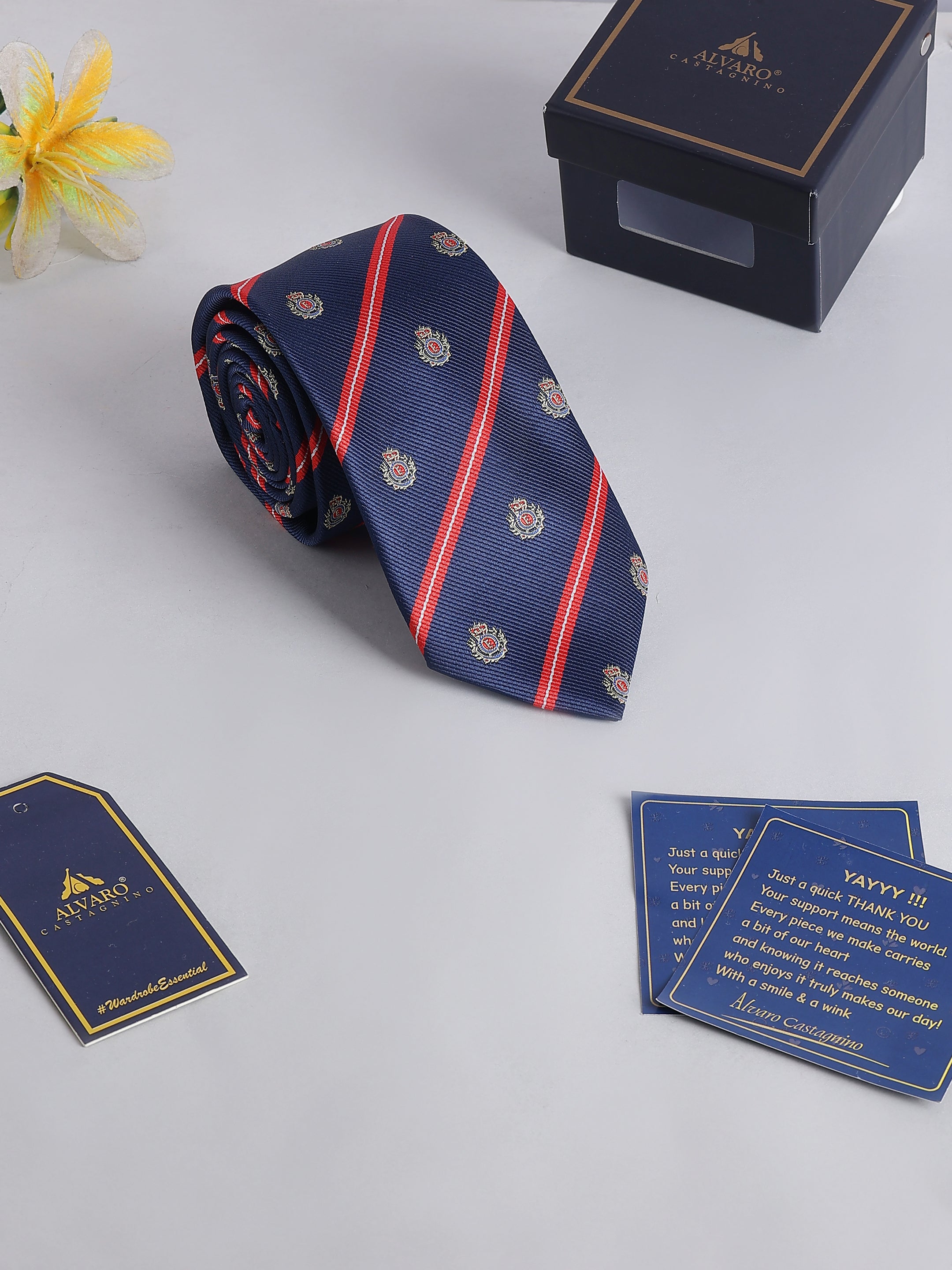 Navy blue tie with red stripes and gold patterns, accompanied by a box and cards on a light gray background.