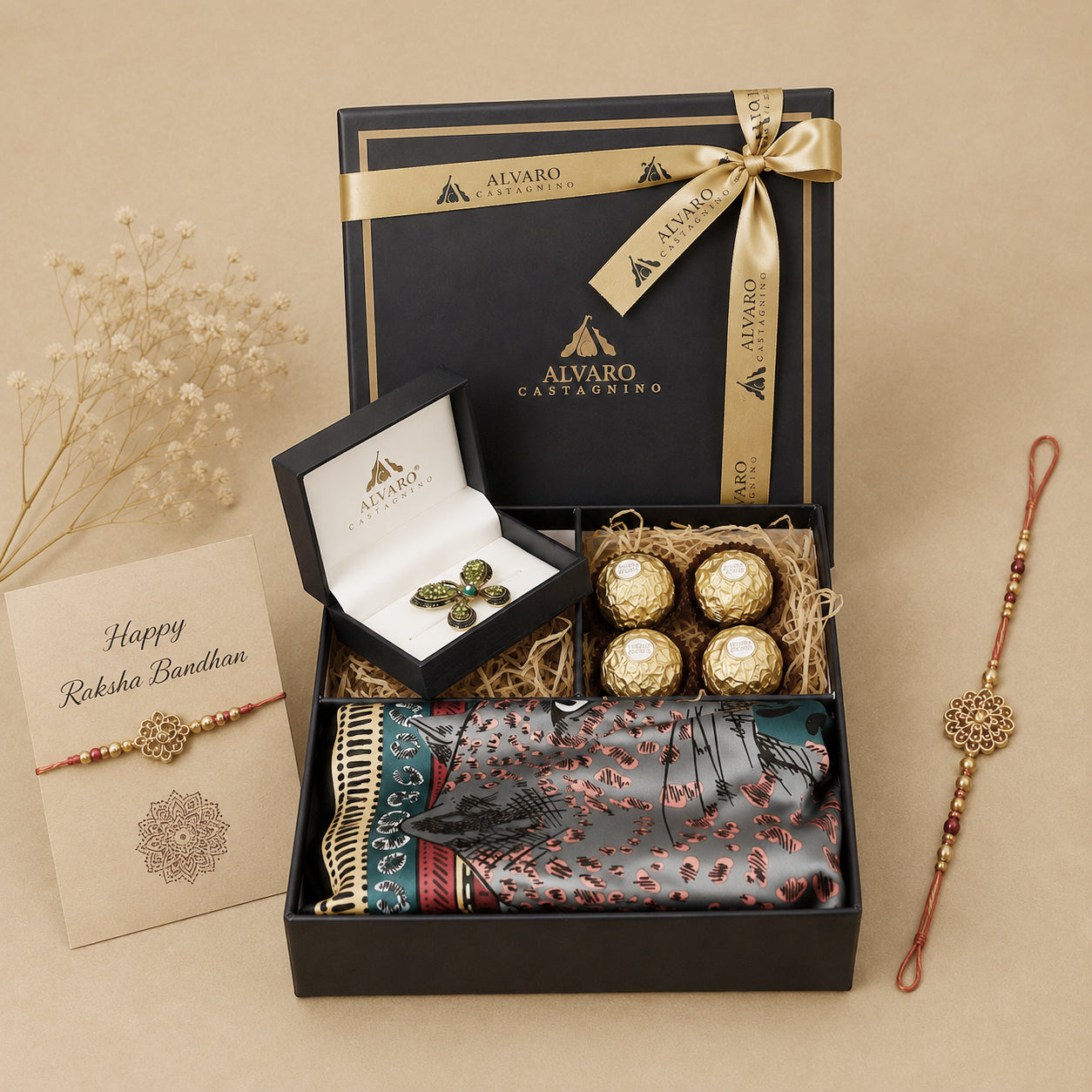 Alvaro Castagnino gift set arranged on a beige background, including an open black presentation box with gold ribbon, a ring box with green stone earrings, four Ferrero Rocher chocolates, a patterned scarf, and two Rakhi bracelets. A greeting card reading Happy Raksha Bandhan is also shown.