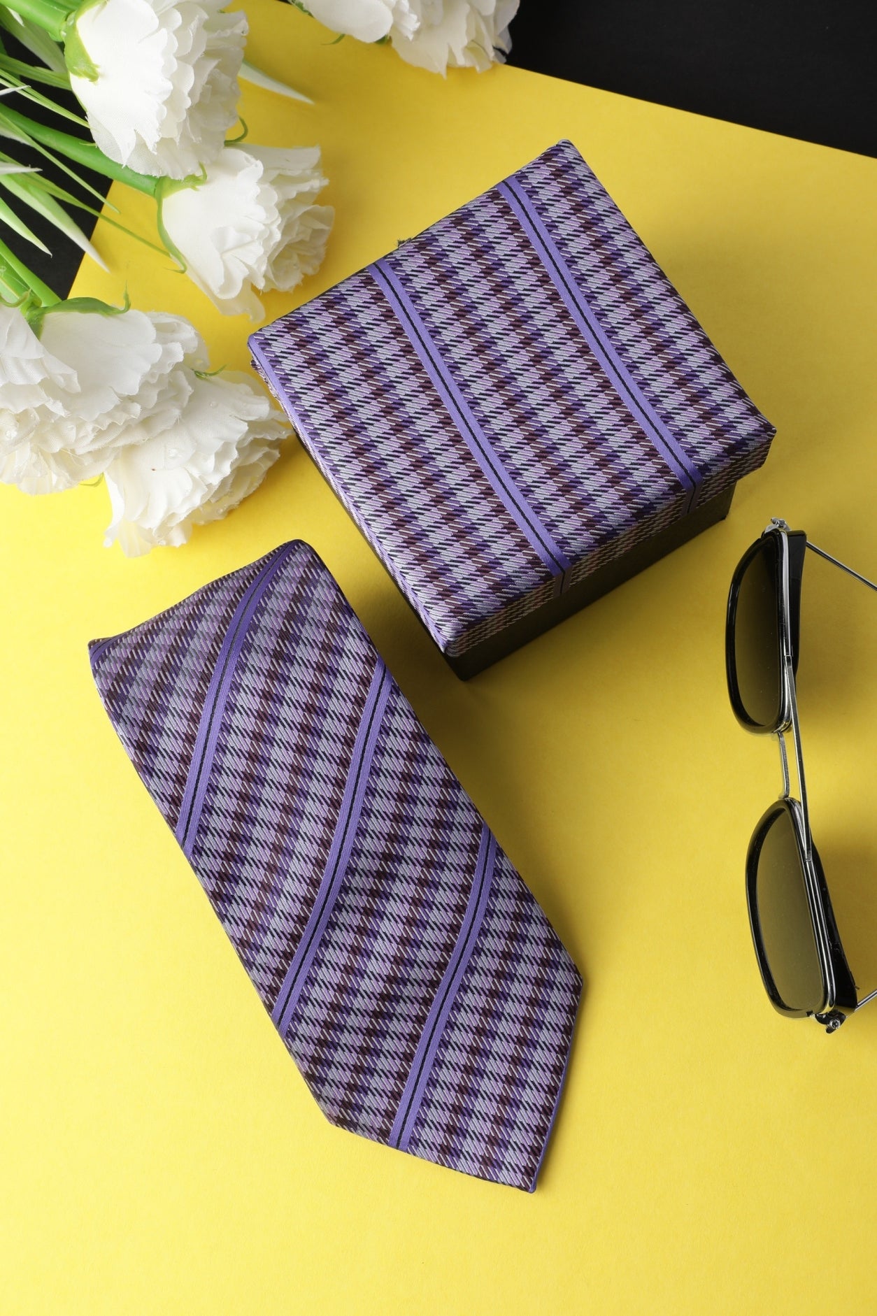 A microfiber necktie with purple and multicolored stripes, displayed next to its luxurious look gift box and a pair of sunglasses, on a yellow background.