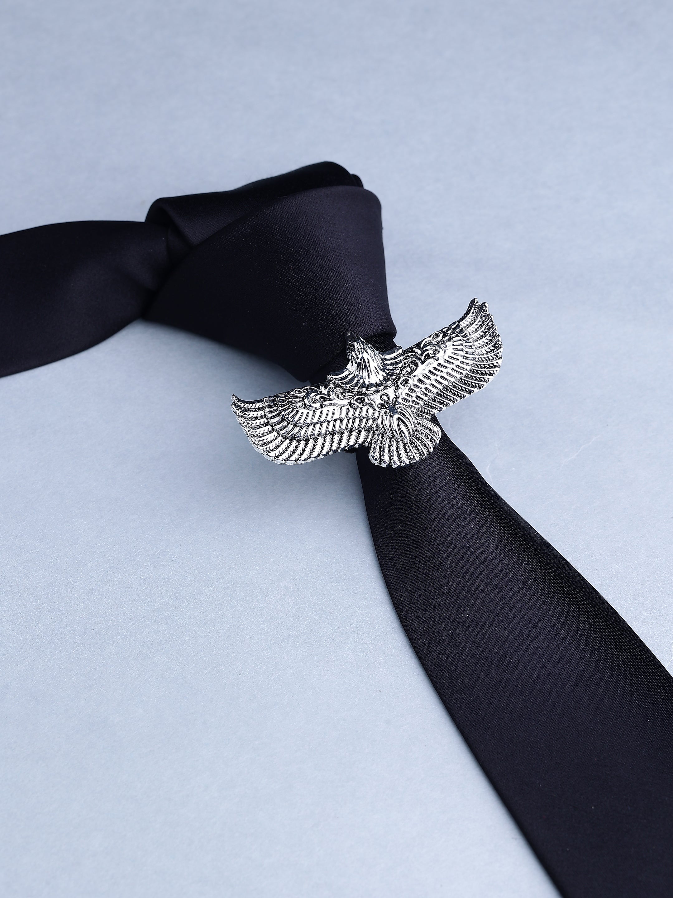 A silver-toned metal tie clip shaped like a detailed eagle with spread wings, holding a black satin necktie against a light blue-gray background.